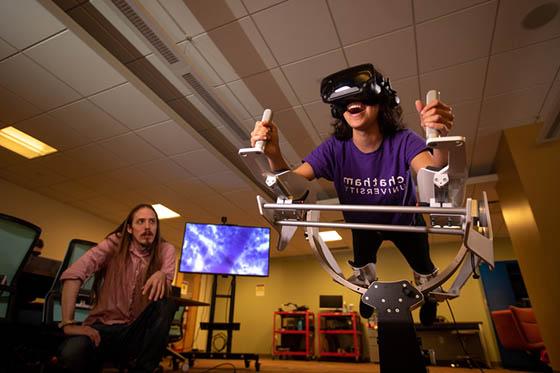 图为一名波胆网站的学生在沉浸式媒体实验室的虚拟现实机器里, while a professor watches